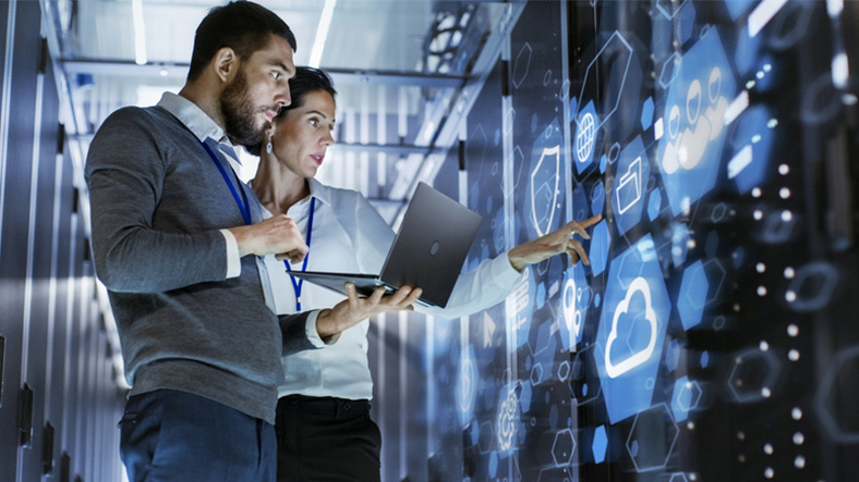 Male IT Specialist Holds Laptop and Discusses Work with Female Server Technician. They're Standing in Data Center, Rack Server Cabinet with Cloud Server Icon and Visualization.