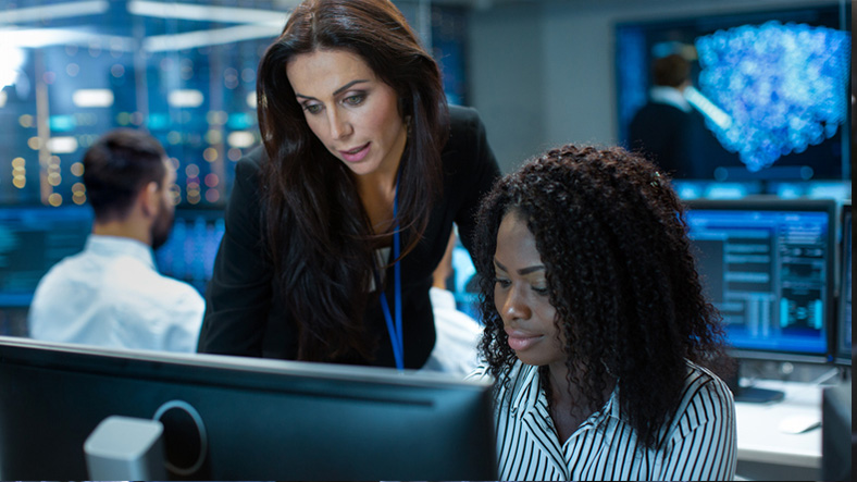 Female Team Leader Consults Young Computer Engineer. They Work in a Crowded Office on a Neural Network/ Artificial Intelligence Project.