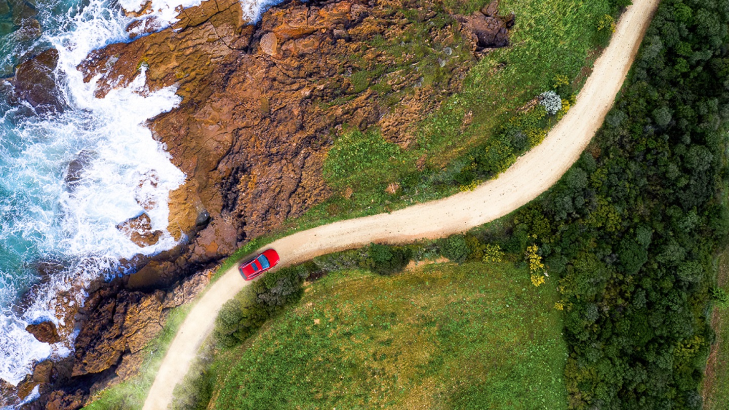 Red car moving on a winding coastal road approaching a beautiful beach with sea waves and blue water. Green forest and rocks for a perfect summer destination. Aerial view.