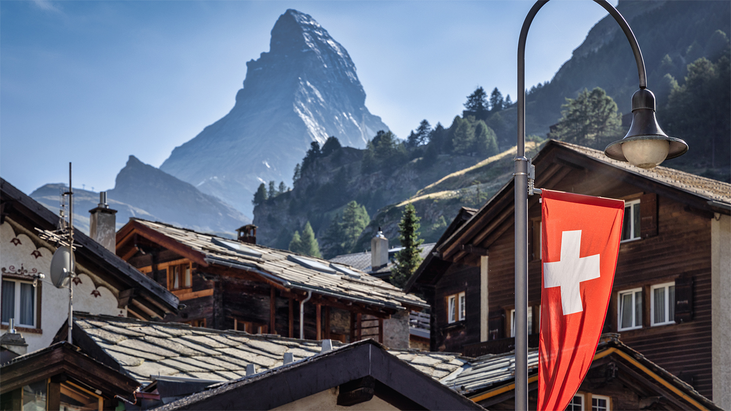 Zermatt Village Typical Traditional Swiss Alps Wooden Houses in the center of Zermatt Village with illuminated Swiss National Flag in the foreground. Famous Matterhorn Mountain Peak under blue summer sky in the background. Zermatt, Valais Canton, Switzerland, Europe