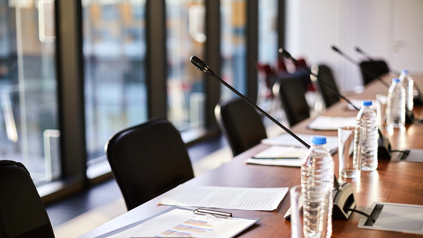 Business documents, plastic bottles of water, glasses and microphones along table and chairs near by in conference hall