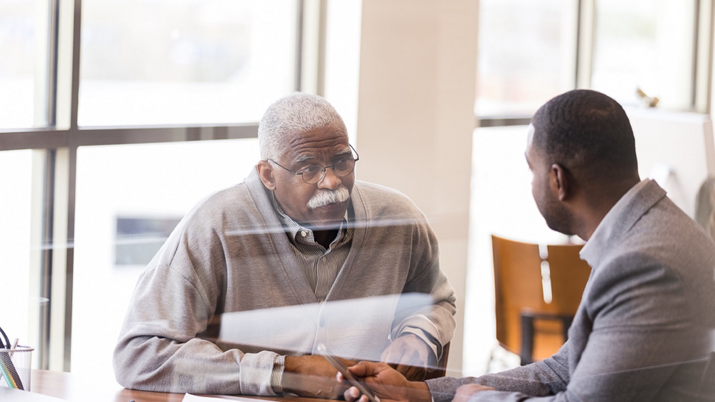 A serious senior man listens attentively as a mid adult male bank employee explains the bank's mobile app.