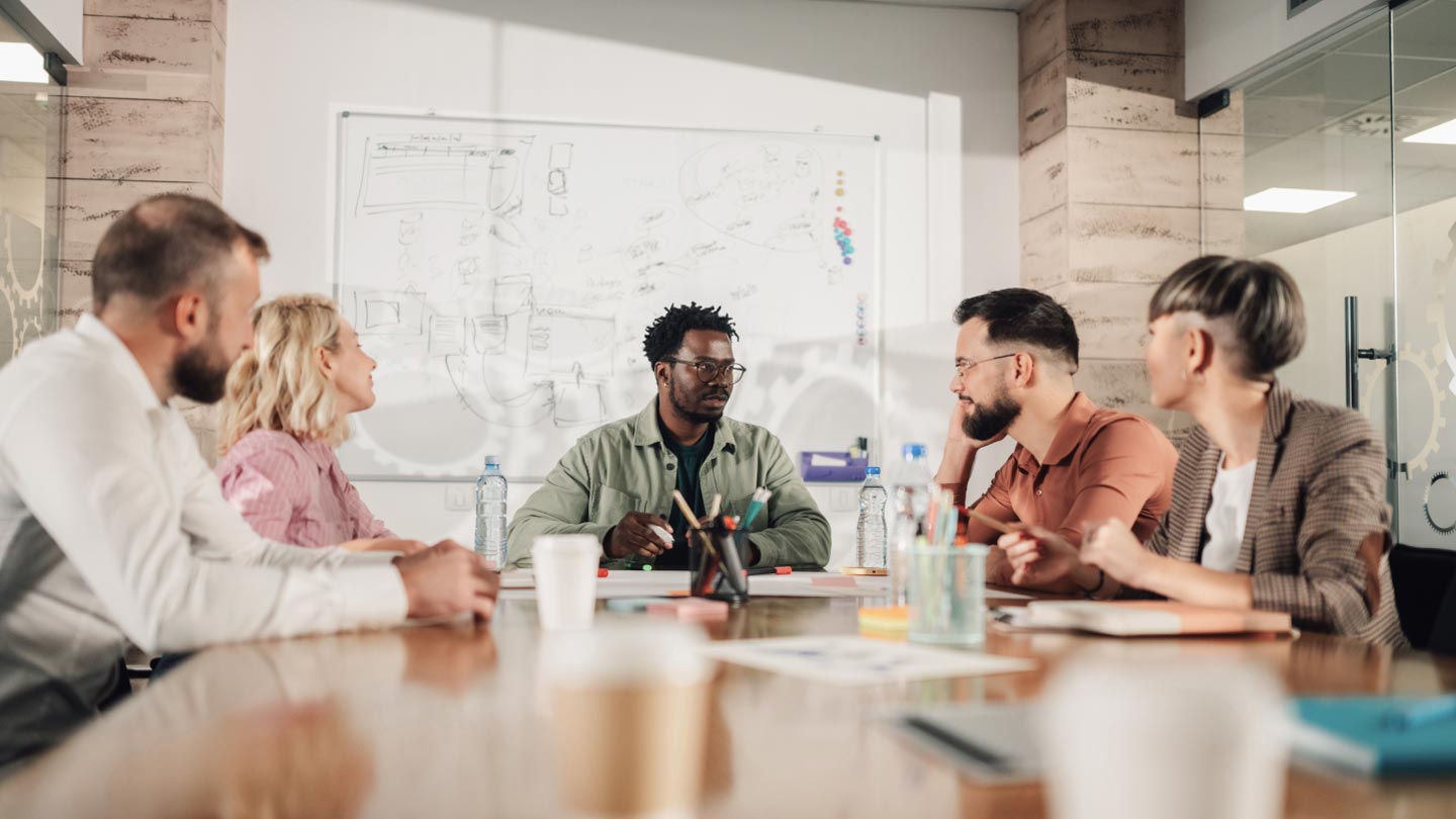 A group of people having a meeting around a conference table with a whiteboard in the background.