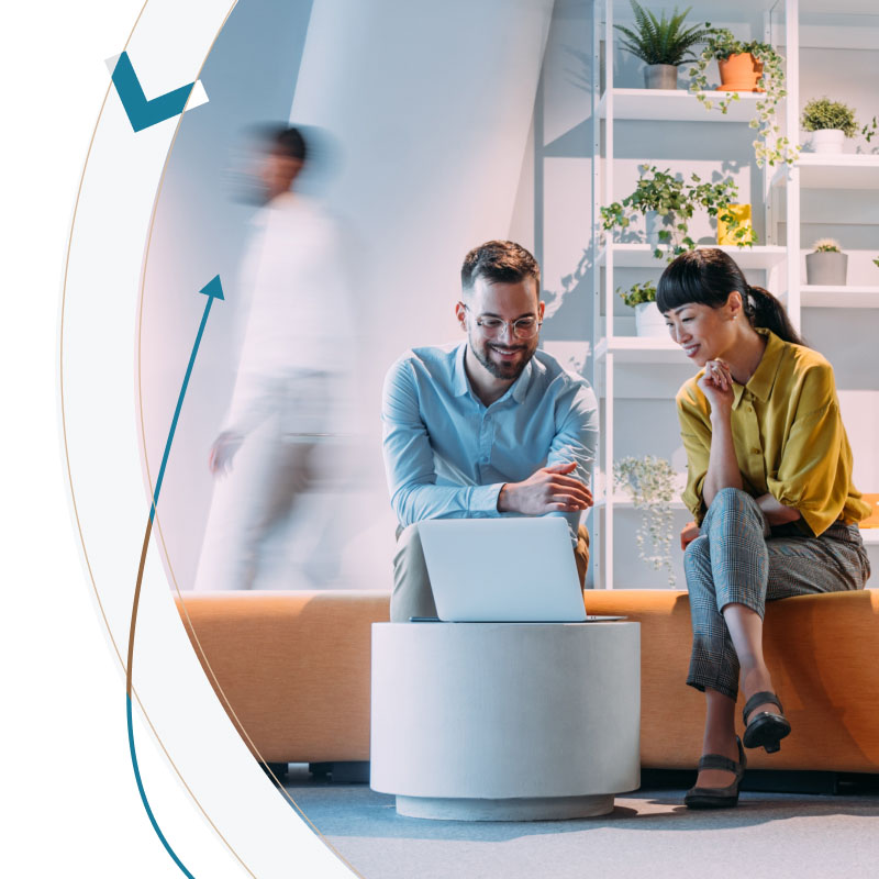 Two people sitting on a modern couch, discussing work with a laptop in a bright office space filled with plants.