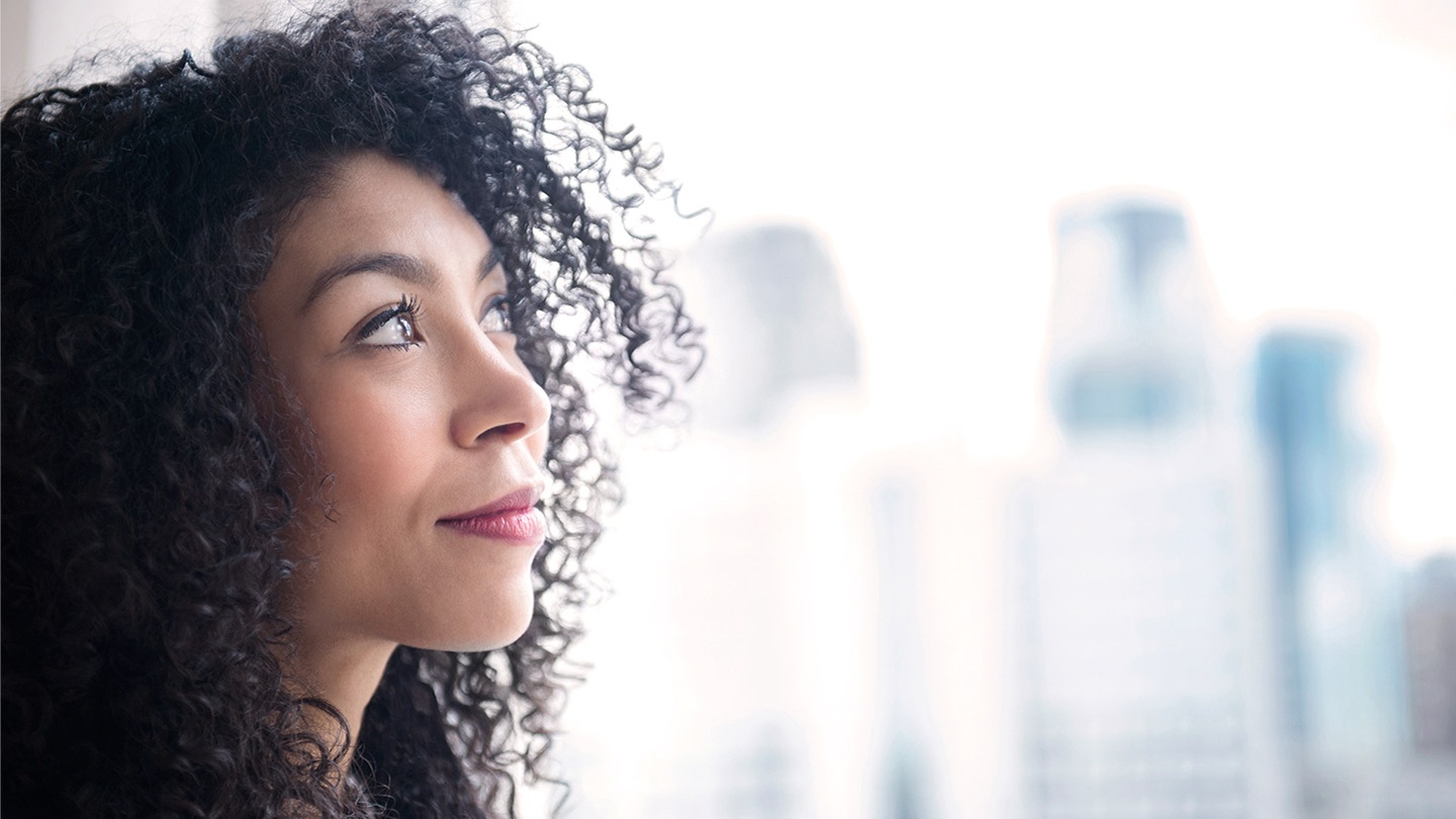 Person with curly hair standing indoors near a window with city buildings in the background.