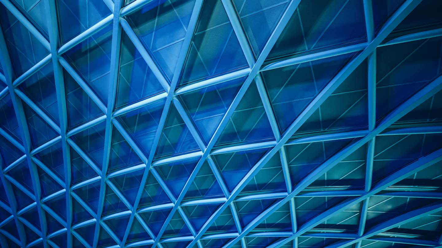 Close-up of a modern glass-and-steel architectural structure with a repeating pattern of blue-tinted triangular panels and intersecting metal beams.
