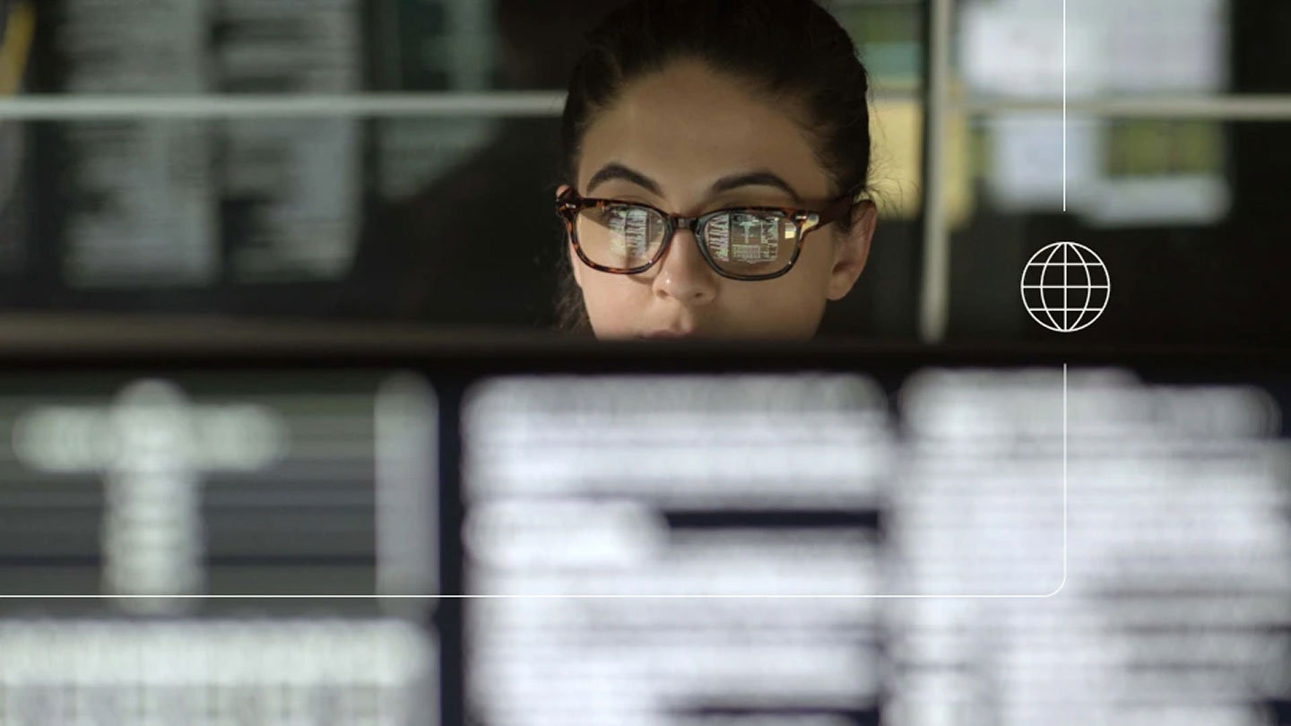 Close-up of a person wearing glasses concentrating on multiple computer screens, with the screen text reflected in the lenses and a small globe icon with thin interface-style overlay lines on the right.
