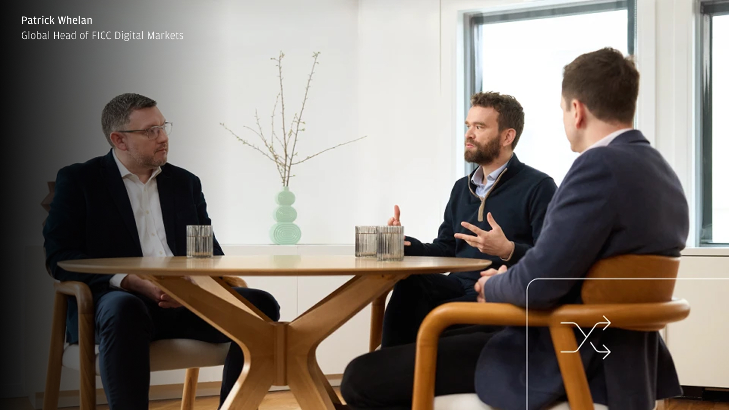 Three people in business attire sit around a wooden table in a bright office, engaged in conversation; one person gestures while speaking, with a name-and-title caption in the top-left and a small interface-style icon overlay in the bottom-right.