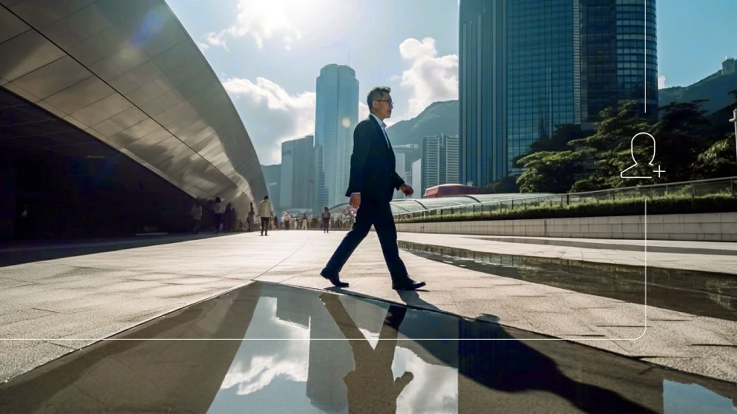 Businessperson in a suit walking across a sunlit modern city plaza, reflected in a shallow pool, with skyscrapers and a curved building in the background and a subtle UI-style person icon overlay.