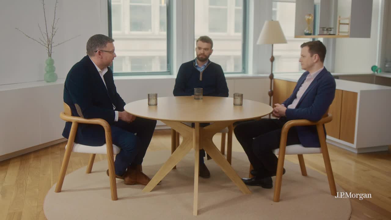 Three people in business attire sit on wooden chairs around a round table in a bright office lounge, with large windows, a floor lamp, and minimal decor in the background; a “J.P. Morgan” watermark appears in the lower-right.