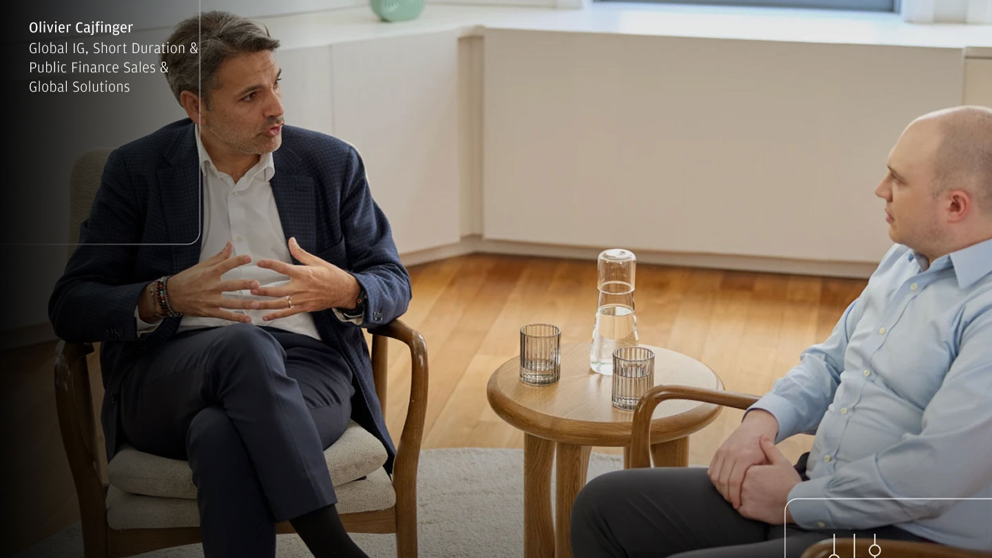 Two people in business attire sit facing each other in a bright office lounge, talking beside a small round table with a water carafe and glasses; a name-and-title text overlay appears in the top-left with subtle UI-style lines/icons.