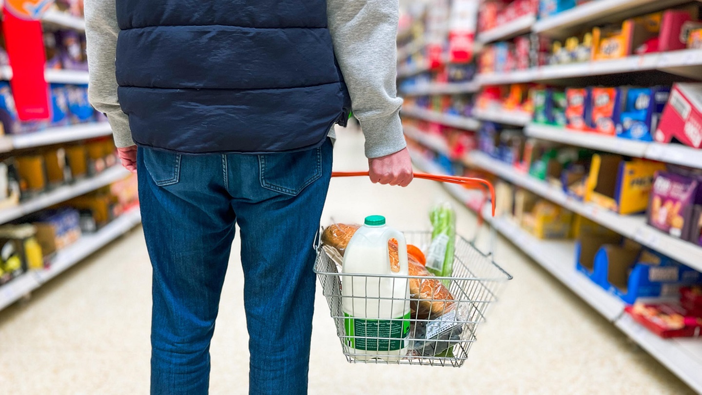 Low angle close up color image depicting a man holding a shopping basked filled with essential fresh groceries like bread and milk in the supermarket.