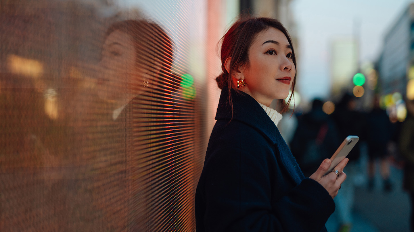 Confident young Asian woman with smartphone, standing against digital display on the street in the city at night. Futuristic concept. Mobile banking concept.