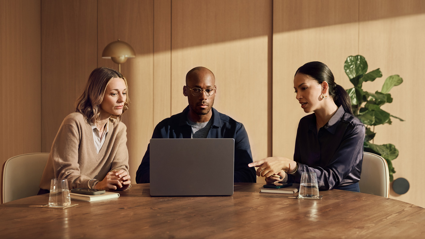 Three people are sitting at a round table in a modern office, engaged in a discussion with a laptop in front of them. banner