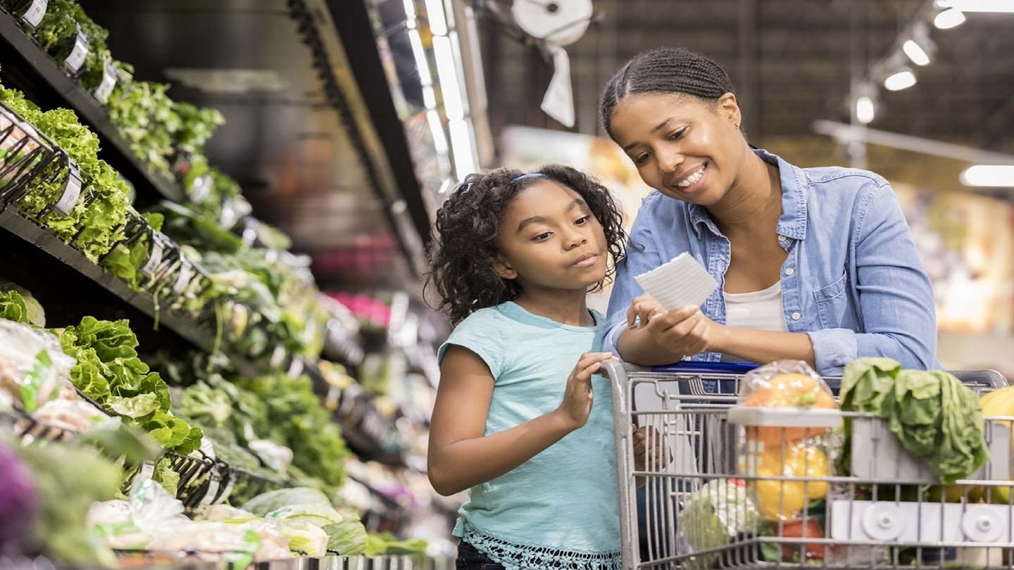 A smiling mid adult woman stands in the produce section of her supermarket and reaches down to show her serious elementary age daughter the next items on a paper shopping list.