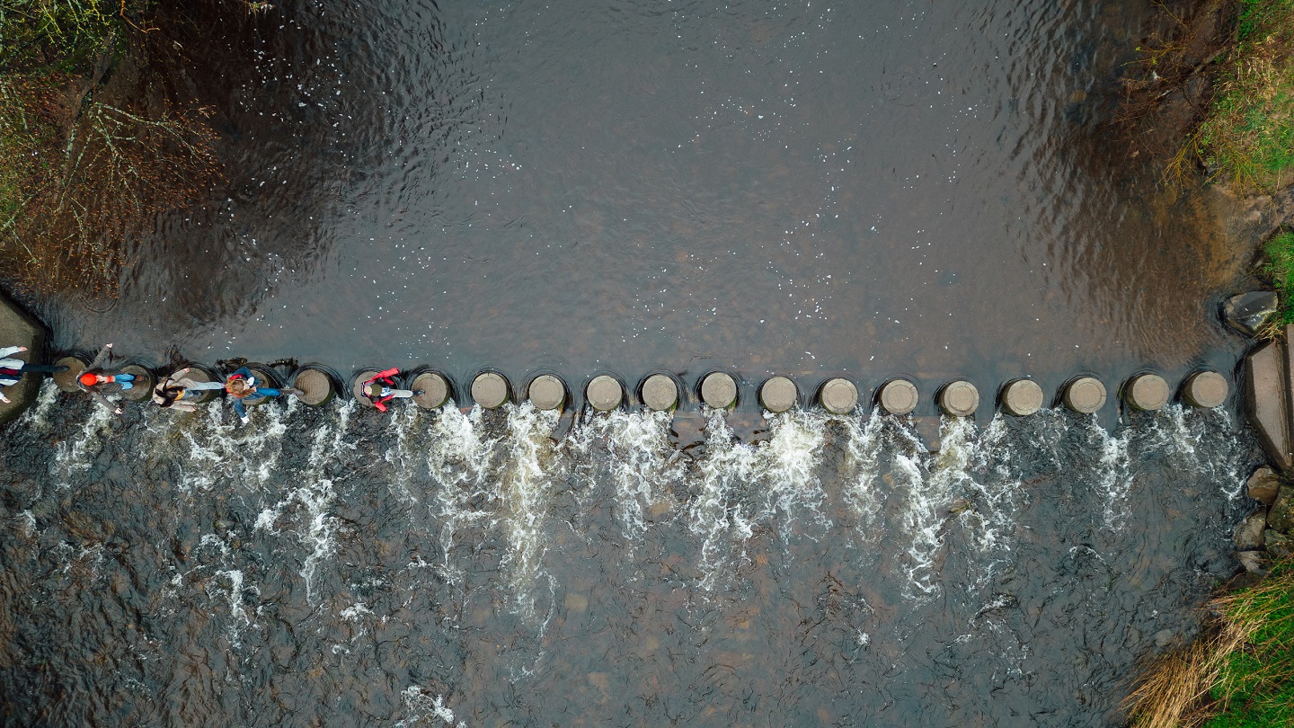 An aerial shot of four students and their teacher wearing warm outdoor clothing on an overcast day in Northumberland. They are on a field trip and are crossing a river by walking over stepping stones.

Video also available of this scenario