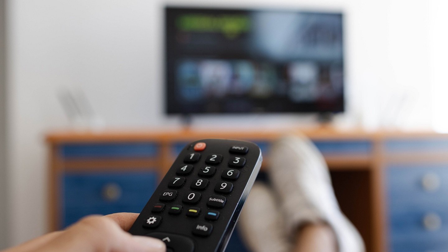 Photo of a hand holding a television remote control while his feet are on the table.