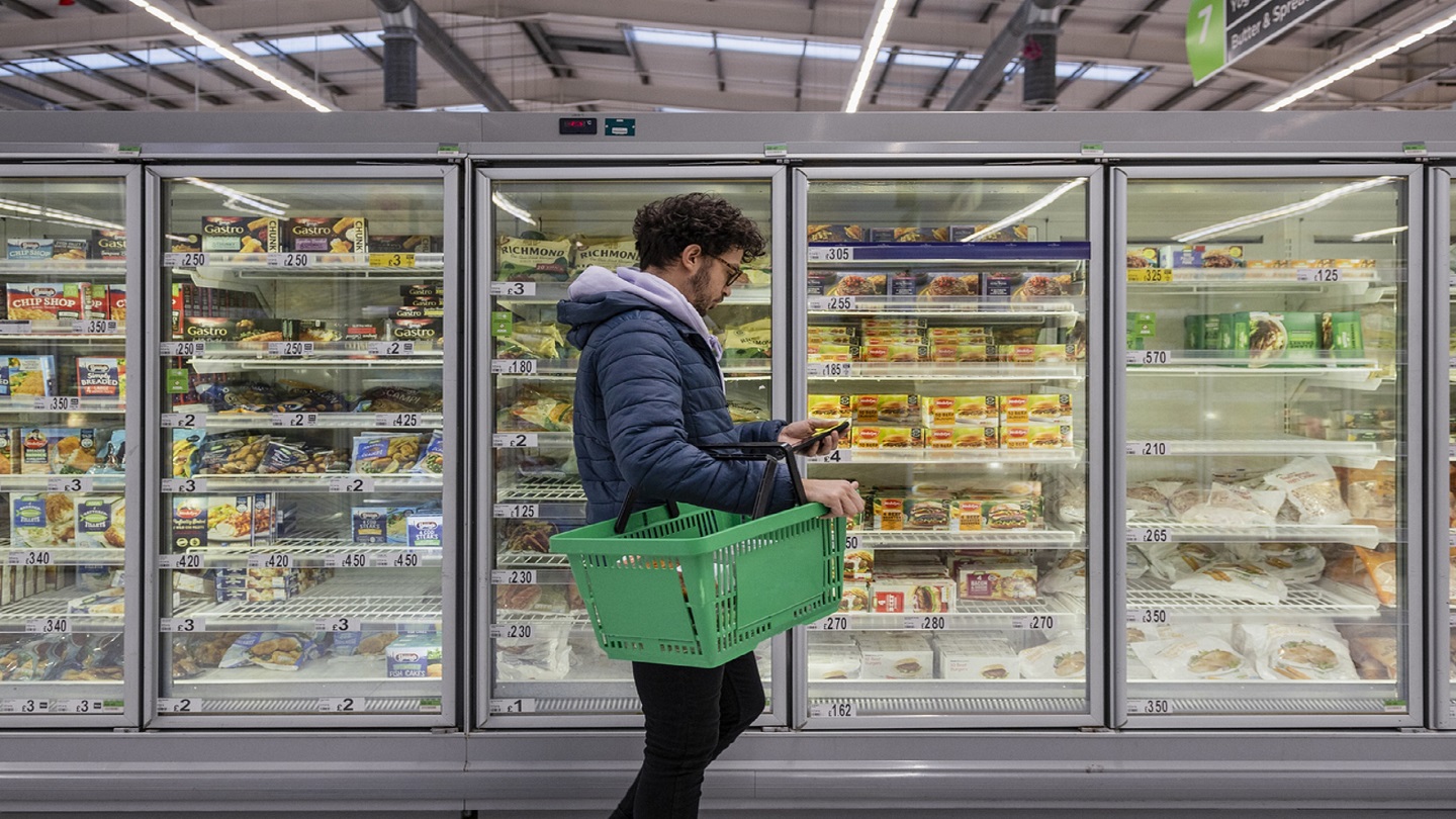 Man shopping in a supermarket while on a budget. He is looking for low prices due to inflation, standing looking at his phone in front of a row of freezers. He is living in the North East of England.