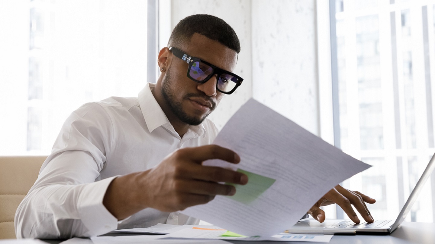 Serious African American manager in stylish glasses reading corporate document with note, sales report, boss order, doing paperwork. Legal expert checking document at workplace with laptop