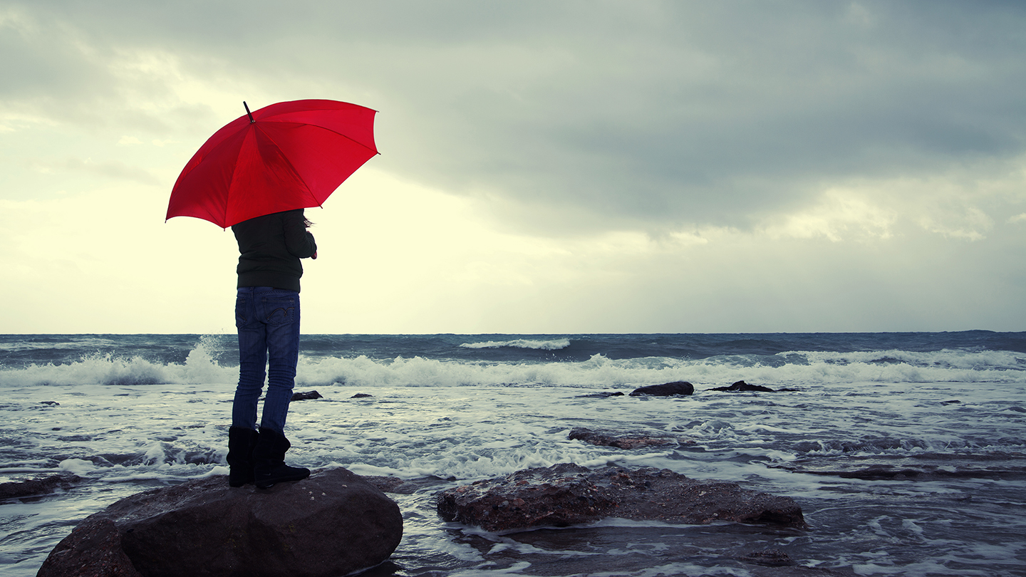 Young girl with red umbrella standing on a stone at the beach and looking away