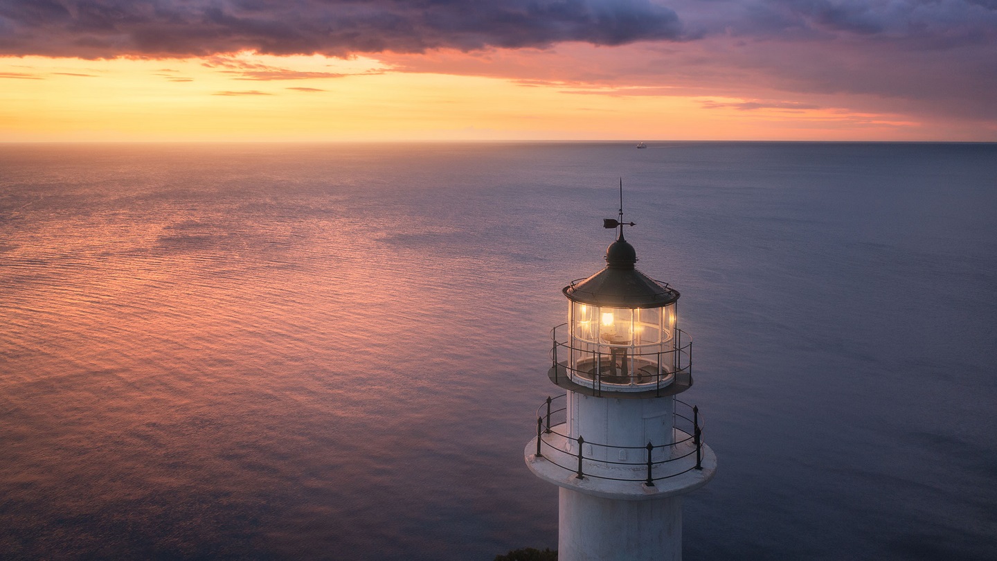 Lighthouse on the mountain peak at colorful sunset in summer. Aerial view. Beautiful lighthouse, light, sea and orange sky with purple clouds at night. Top view of Cape Lefkada, Greece. Landscape